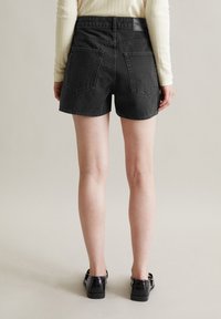 Zwarte denim shorts met een hoge taille en opgerolde zoom, voorzien van twee achterzakken en een gestructureerde tailleband. Draagt een paar zwarte schoenen.