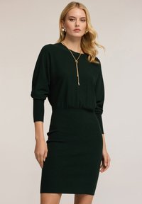 Abito lavorato a maglia verde scuro con silhouette aderente, maniche a tre quarti e vita segnala. La modella indossa una lunga collana dorata e orecchini.
