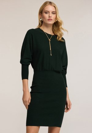 Abito lavorato a maglia verde scuro con silhouette aderente, maniche a tre quarti e vita segnala. La modella indossa una lunga collana dorata e orecchini.