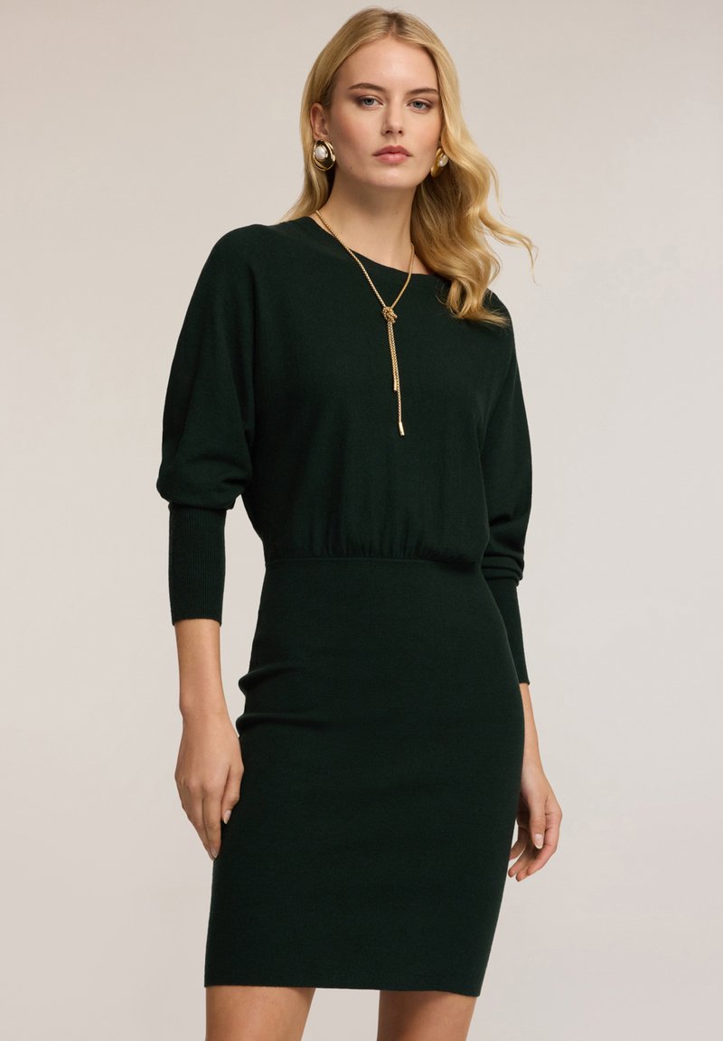 Abito lavorato a maglia verde scuro con silhouette aderente, maniche a tre quarti e vita segnala. La modella indossa una lunga collana dorata e orecchini.