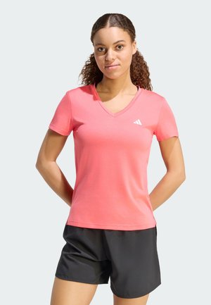 Joven con cabello rizado que lleva una camiseta rosa de cuello en V y pantalones cortos deportivos negros, de pie con las manos detrás de la espalda.