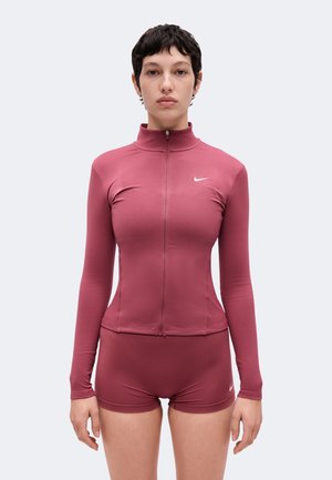 Femme portant un haut moulant mauve à manches longues avec fermeture éclair et un short assorti avec des logos Nike blancs, en position face.