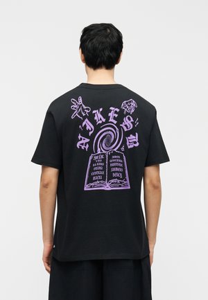 Osoba w czarnej koszulce z fioletowym nadrukiem przedstawiającym dłonie, wirujący spiralny wzór oraz otwartą książkę z tekstem na plecach.