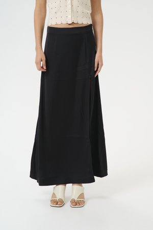 Black maxi skirt with a smooth texture, side slit, elastic waistband, and a relaxed fit, paired with beige open-toe sandals.