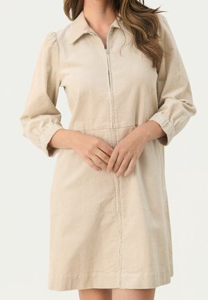 Beżowa sukienka ze sztruksu z rękawami 3/4 o kroju bufek, suwakiem z przodu i kołnierzykiem, noszona przez osobę z długimi włosami, z rękami złożonymi na wysokości talii.