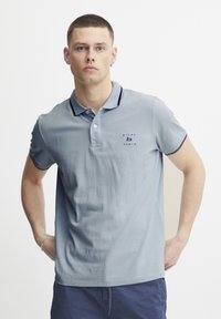 Lys blå polo-skjorte med krage og tre knapper. Har en marineblå aksent på kragen og en liten logo på brystet. Glatt tekstur.