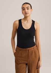 Svart ribbad linne med rundad halsringning, kombinerat med bruna veckade byxor som har dragsko i midjan och avslappnad passform. Silverhalsband som accent.