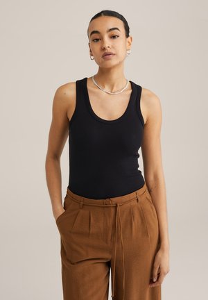 Schwarzes geripptes Tanktop mit rundem Ausschnitt, kombiniert mit braunen, plissierten Hosen, die einen Kordelzug an der Taille und eine lockere Passform haben. Akzentuiertes silbernes Halskettchen.