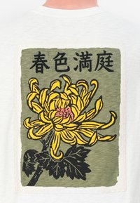 White t-shirt featuring a large, embroidered yellow chrysanthemum design on a green rectangular background, framed with a textured border.