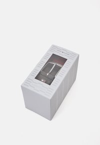 Caja de embalaje gris con textura acanalada vertical, ventana transparente que muestra una hebilla de cinturón plateada y una correa de cuero negro, con acentos del logo.
