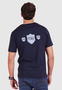 T-shirt en coton bleu marine à manches courtes, avec un motif en forme de bouclier portant "Ruckfield Club" et la date 1977 brodés en blanc et gris au dos.