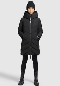 Zwarte pufferjas met een hoge kraag, chevronpatroon, voorrits en zijzakken. Gepaard met zwarte leggings en enkellaarsjes.