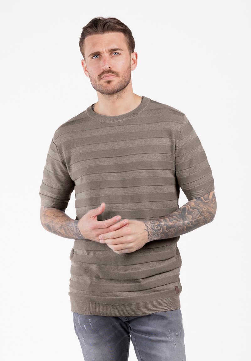 Uomo con tatuaggi che indossa un maglione a strisce taupe a maniche corte e jeans grigi strappati, in piedi su uno sfondo bianco.
