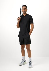 Camisa polo preta de mangas curtas com colarinho, calções pretos, meias brancas e sapatilhas desportivas azul claro. A segurar uma raquete de ténis preta.