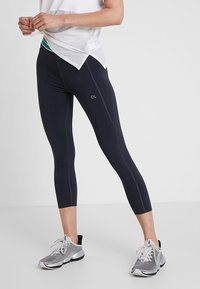 Vrouw in zwarte cropped leggings, witte mesh top en grijze sportschoenen, staand met gespreide benen tegen een witte achtergrond.