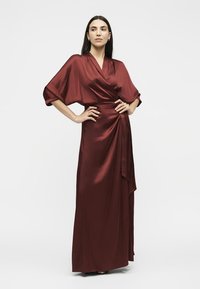 Femme debout, les mains sur les hanches, portant une longue robe portefeuille en satin bordeaux avec des manches larges sur fond blanc.