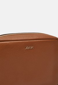 Pochette en cuir brun avec une surface texturée, fermeture éclair dorée et logo "JOOP!" en relief sur le devant. Forme rectangulaire, design sophistiqué.
