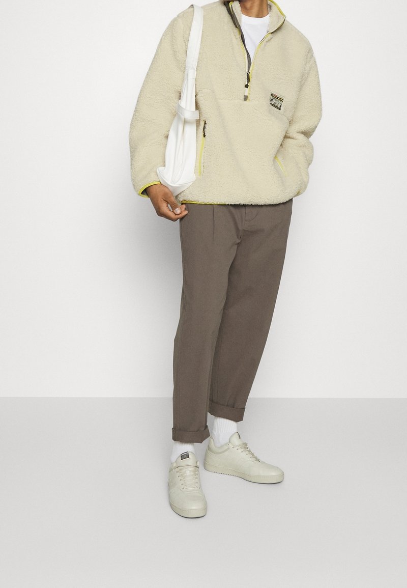 Chaqueta de forro polar color crema con media cremallera, ribetes amarillos en contraste, pantalones sueltos marrones y zapatillas blancas con detalles texturizados.
