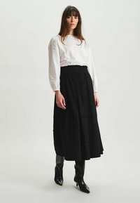 Un haut blanc à manches longues associé à une jupe maxi noire à volants. La tenue est complétée par des bottes noires brillantes jusqu'aux genoux.