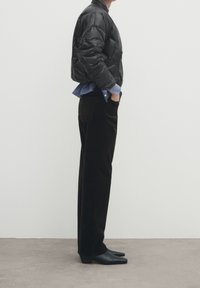 Person iført sort cropped pufferjakke, blå skjorte, sorte bukser og sorte spidse lædersko, der står sidelæns på grå gulv.