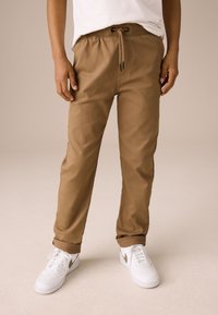 Pantaloni maro, ușori, din amestec de bumbac, cu talie elastică și șnur. Manșete rulate și buzunare vizibile. Asortați cu adidași albi.