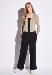 Ljusbeige, strukturerad kofta över en svart topp, matchad med svarta vida byxor och vita sneakers på en modell mot en vit bakgrund.