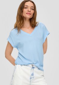Lichtblauwe, kortemouw V-hals top van textiel met een gestructureerde stof. Heeft een ontspannen pasvorm en opgerolde mouwmanchetten. Gepaarde met witte denim shorts.
