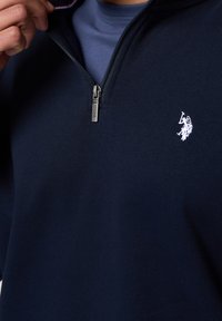 Mörkblå zip-up-tröja i mjukt tyg; har en liten vit broderad logga och kontrasterande foder på insidan av kragen.