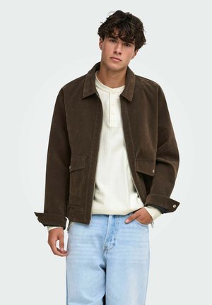 Jeune homme aux cheveux bouclés portant une veste marron foncé par-dessus une chemise blanche à manches longues et un jean bleu clair, debout devant un fond uni.