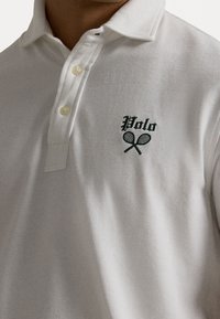 Bílá polokošile vyrobená z texturované látky, s vyšitým zeleným logem "Polo" a zkříženými tenisovými raketami na hrudi. Tříknoflíkový zapínání.