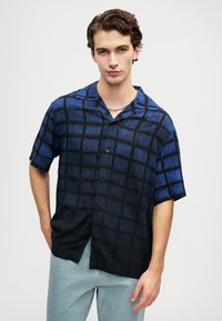 Ung mann iført en blå og svart rutete kortermet skjorte, lyseblå denimbukser og et sølvkjede, med avslappede hender.