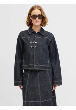 Kobieta z krótkimi falowanymi włosami, nosząca duże czarne okulary przeciwsłoneczne, ciemną dżinsową kurtkę z dwoma metalowymi klamrami i dopasowaną spódnicę.