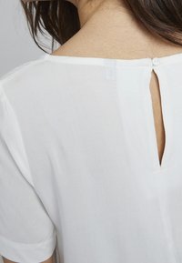 Vit blus med en nyckelhålsöppning bak, korta ärmar och mjukt, lättviktigt tyg. Sömdetalj synlig nära ringningen.