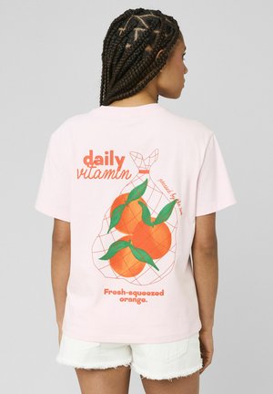 Jeune personne aux cheveux tressés portant un t-shirt rose clair avec trois oranges en filet et un texte sur la vitamine quotidienne et l'orange fraîchement pressée.