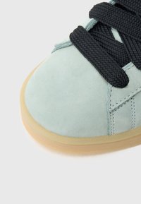 Αθλητικό παπούτσι από γαλάζιο σουέντ με μαύρα υφασμένα κορδόνια, στρογγυλή μύτη και σόλα από καουτσούκ γκαμ· διαθέτει ραφές κατά μήκος της πλευράς.