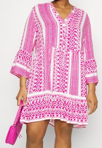 Vestido rosa con escote en V y mangas acampanadas, que presenta patrones geométricos en rayas alternas. Un dobladillo con volantes añade detalle. Acompañado de un bolso rosa.