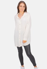 Woman standing in white buttoned long-sleeve shirt, black leather-look leggings, and white sneakers against a plain background.