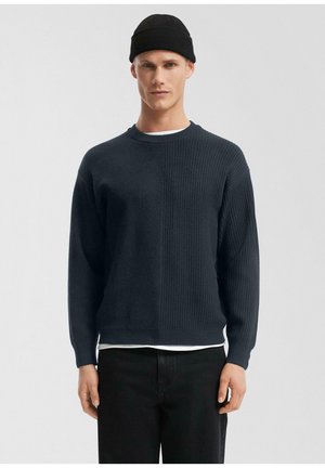 Młody mężczyzna w czarnej czapce z daszkiem, granatowym prążkowanym swetrze na białej koszuli i czarnych spodniach, stojący na jednolitym jasnoszarym tle.