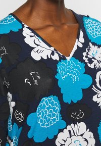 Granatowa bluzka z dekoltem w serek, ozdobiona kwiatowym wzorem w odcieniach turkusu, czerni i bieli. Miękki materiał o gładkiej fakturze, podkreślający różnorodne kształty kwiatów.