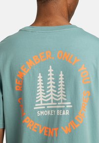 Camiseta de algodón en verde claro con texto en naranja y blanco: "RECUERDA, SOLO TÚ PUEDES PREVENIR INCENDIOS FORESTALES" con un gráfico de árbol. Mangas cortas.