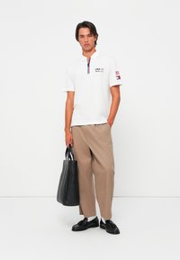 Hvid polo shirt med lynlås, marineblå og røde detaljer, prydet med "USA" og flag broderi. Sammen med beige bukser og en sort tote bag.