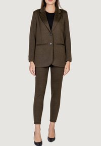 Blazer de espiga marrón con dos botones y bolsillos, combinado con pantalones ajustados a juego. Llevado con tacones negros, tejido texturizado.