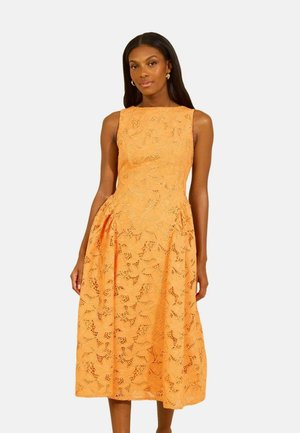 Kvinde iført ærmeløs orange midikjole med blomstermønster, stående foran en ensfarvet lys baggrund.