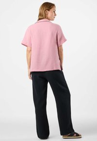 Chemise à manches courtes en tissu rose texturé, associée à un pantalon noir large. Elle possède un col et des fentes sur les côtés à l'ourlet. Coupe décontractée.
