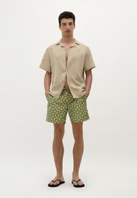 Cămașă scurtă, de culoare bej, pantaloni scurți de baie cu model verde și sandale negre cu vârf deschis. Pantalonii scurți de baie au forme geometrice în nuanțe de verde deschis și închis.