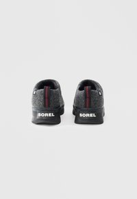 Grå sneakers med svarte gummisåler, med en rød baklapp og hvit "SOREL"-merking på hælen. Rund design.