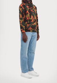 Sudadera con capucha que presenta un patrón de camuflaje oscuro con acentos naranjas, combinada con unos vaqueros de color azul claro y zapatillas blancas.