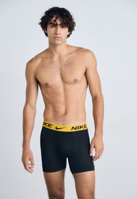 Boxers noirs avec un logo Nike jaune sur la ceinture, fabriqués dans un matériau lisse et extensible. Le modèle se tient avec les bras détendus.