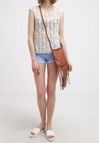 Top in pizzo con motivo floreale, colore beige chiaro; abbinato a pantaloni shorts in denim azzurro. Borsa a tracolla con frange marrone. Sandali bianchi a scivolo.