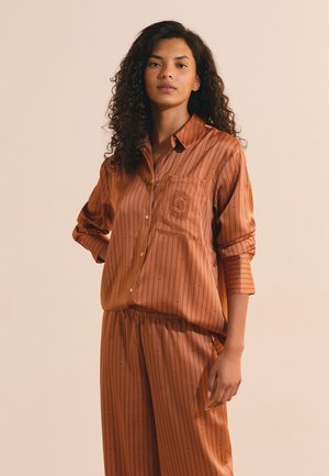 Woman with curly hair wearing a rust-colored striped button-up shirt with matching loose pants, standing against a plain beige background.